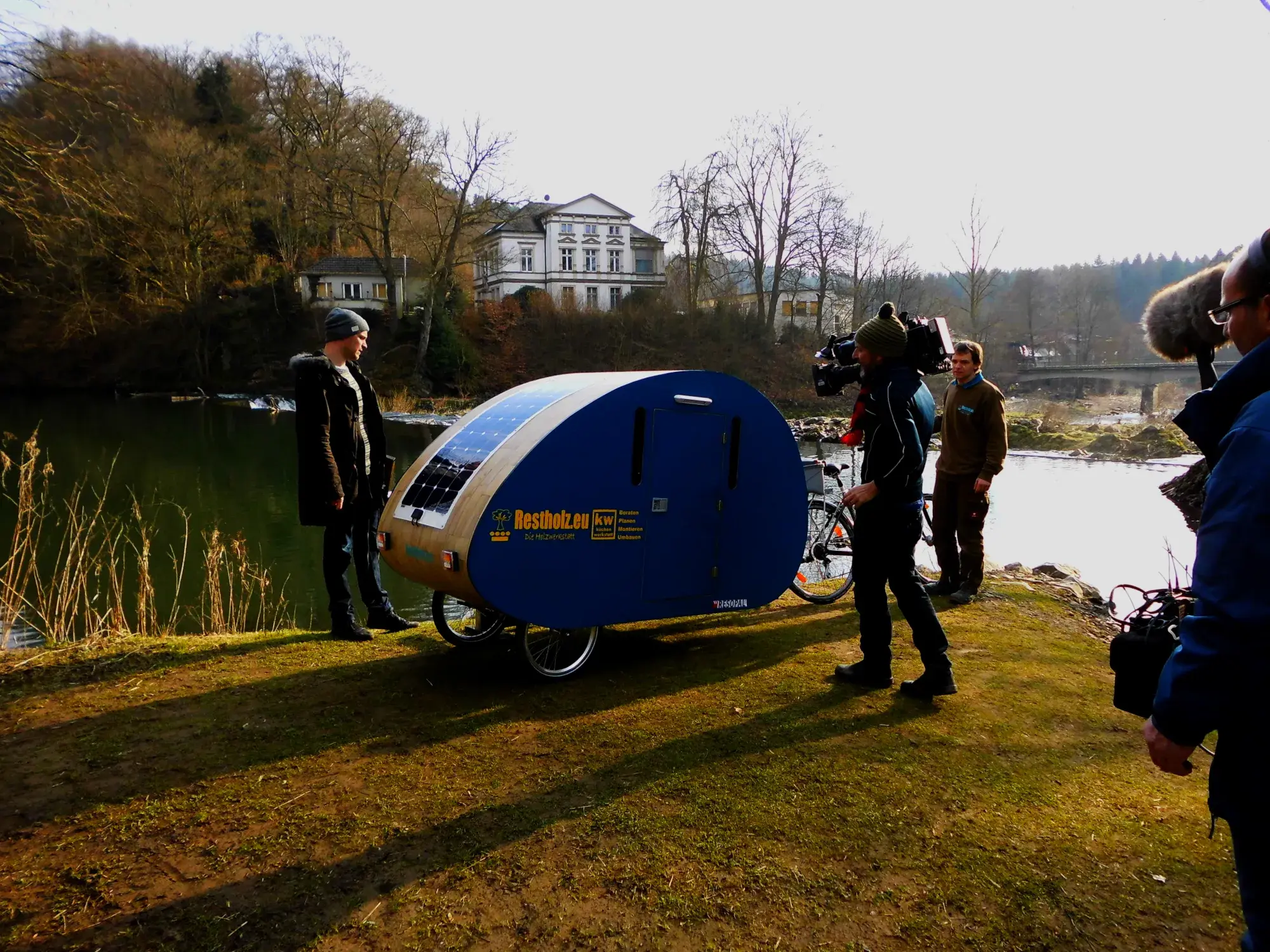 Mann mit Mini-Wohnwagen am See, umgeben von Kamerateam, Bäume im Hintergrund