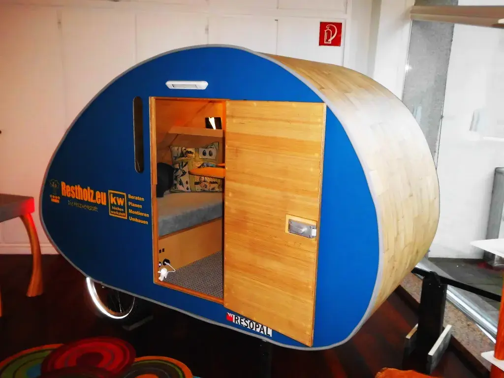 Kleiner, blauer Wohnwagen mit Holztür und Fenster steht im Raum.